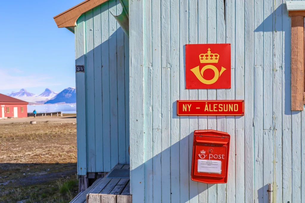Röd postlåda med skylten NY-ÅLESUND på en ljusblå träfasad