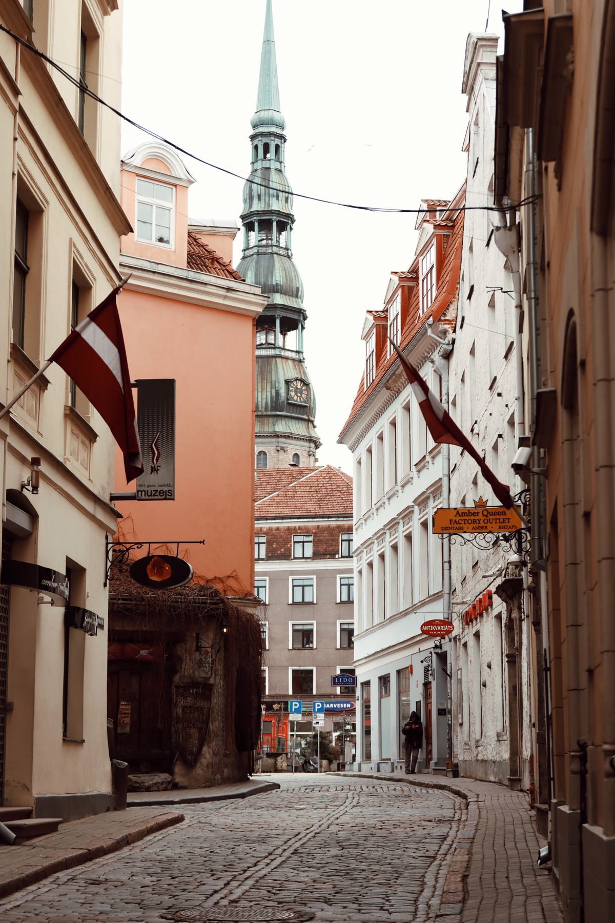 Kullerstensgata i Rigas gamla stad med lettiska flaggor och Sankt Petrikyrkans torn